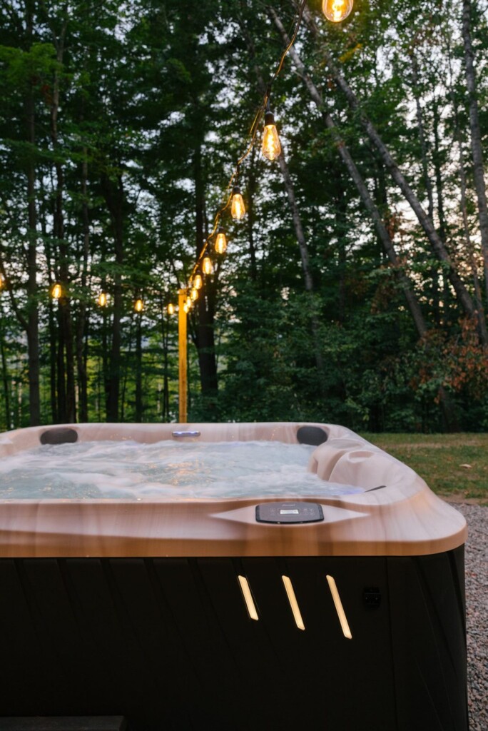 Outdoor hot tub glowing at dusk under string lights in the wooded backyard
